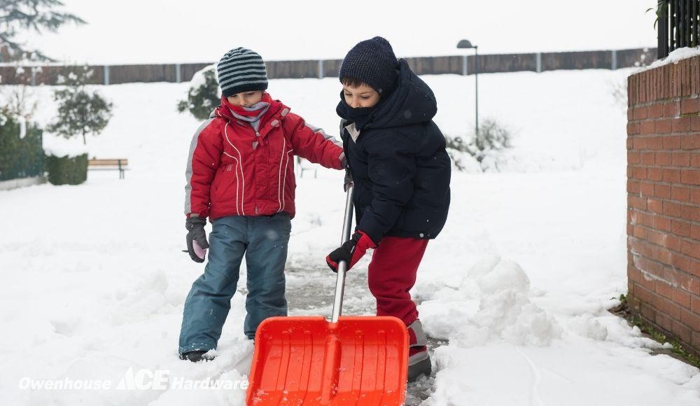 Two children using a red snow shovel to clear snow from a walkway.